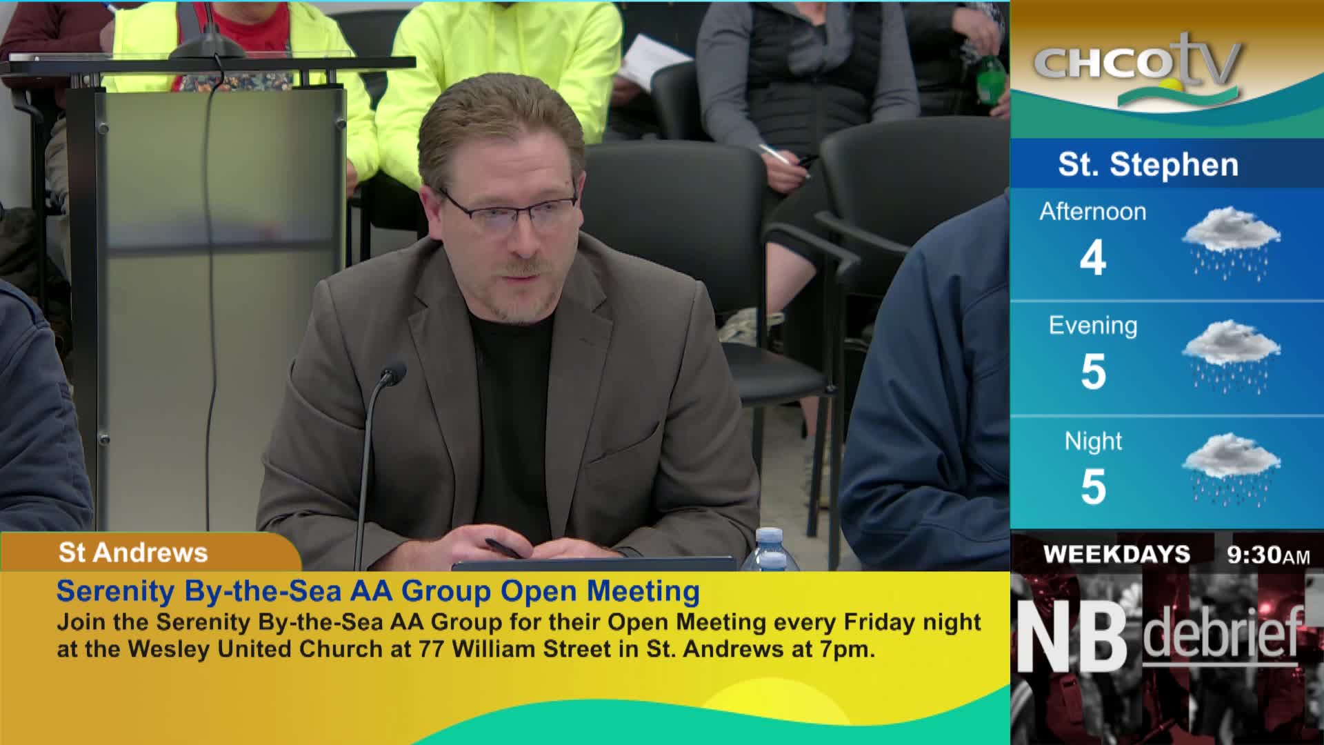 A man in a suit speaks into a microphone. To his right, a weather report for St. Stephen shows rain throughout the day. A man in a suit speaks into a microphone. To his right, a weather report for St. Stephen shows rain throughout the day.