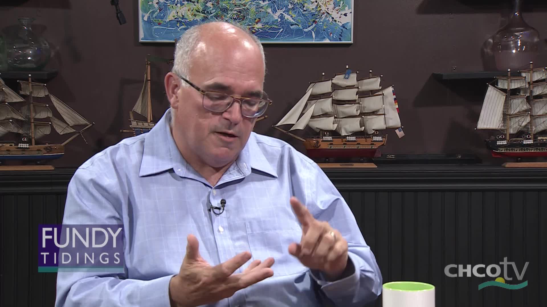 A man in a blue shirt gestures with his hands as he speaks. Behind him, model ships sit on a shelf, and the "Fundy Tidings" logo appears on screen. A man in a blue shirt gestures with his hands as he speaks. Behind him, model ships sit on a shelf, and the "Fundy Tidings" logo appears on screen.