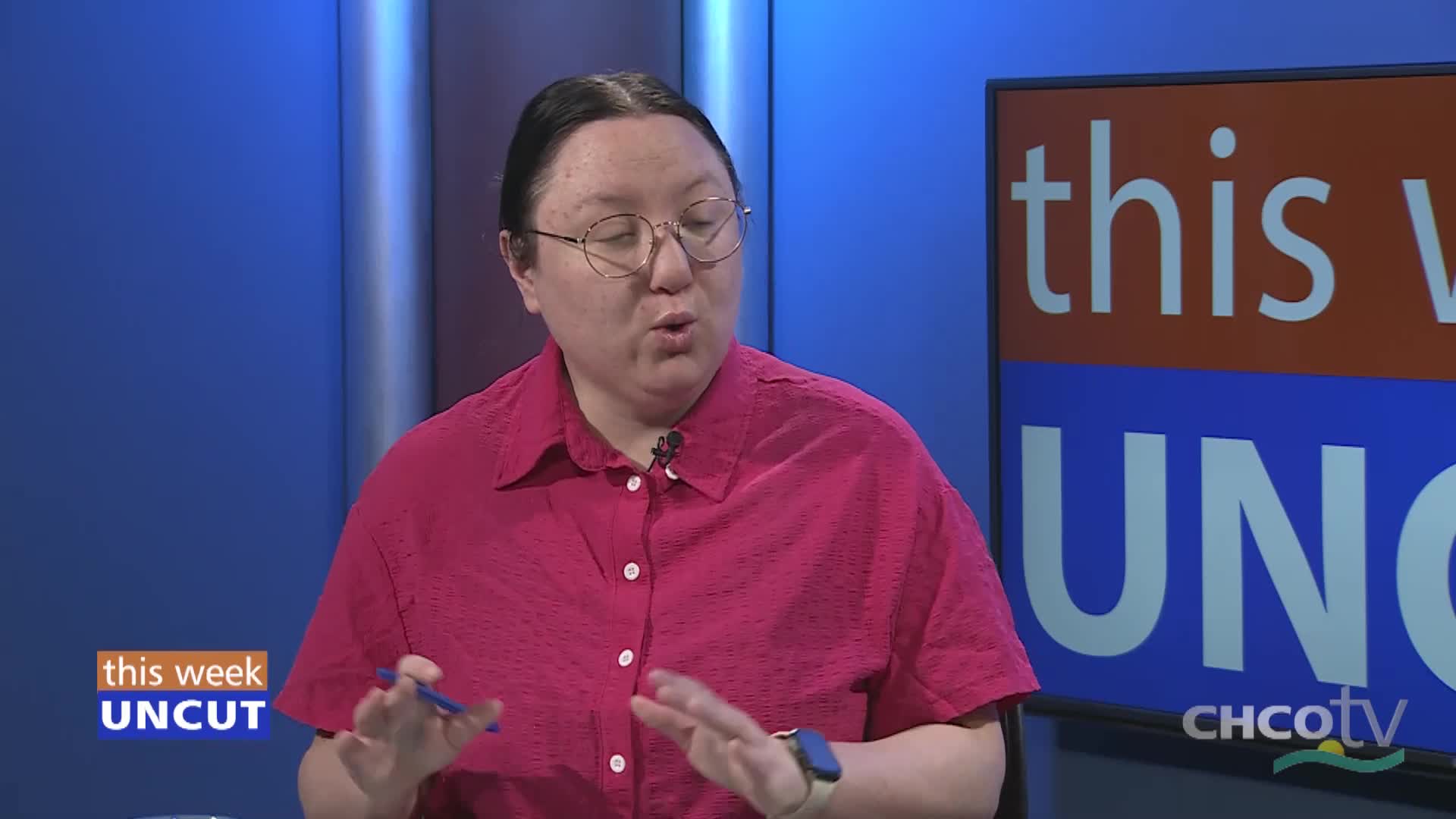 A woman in a bright pink shirt speaks animatedly, gesturing with a blue pen. Behind her, a screen displays "this week UNCUT" with the CHCO-TV logo.