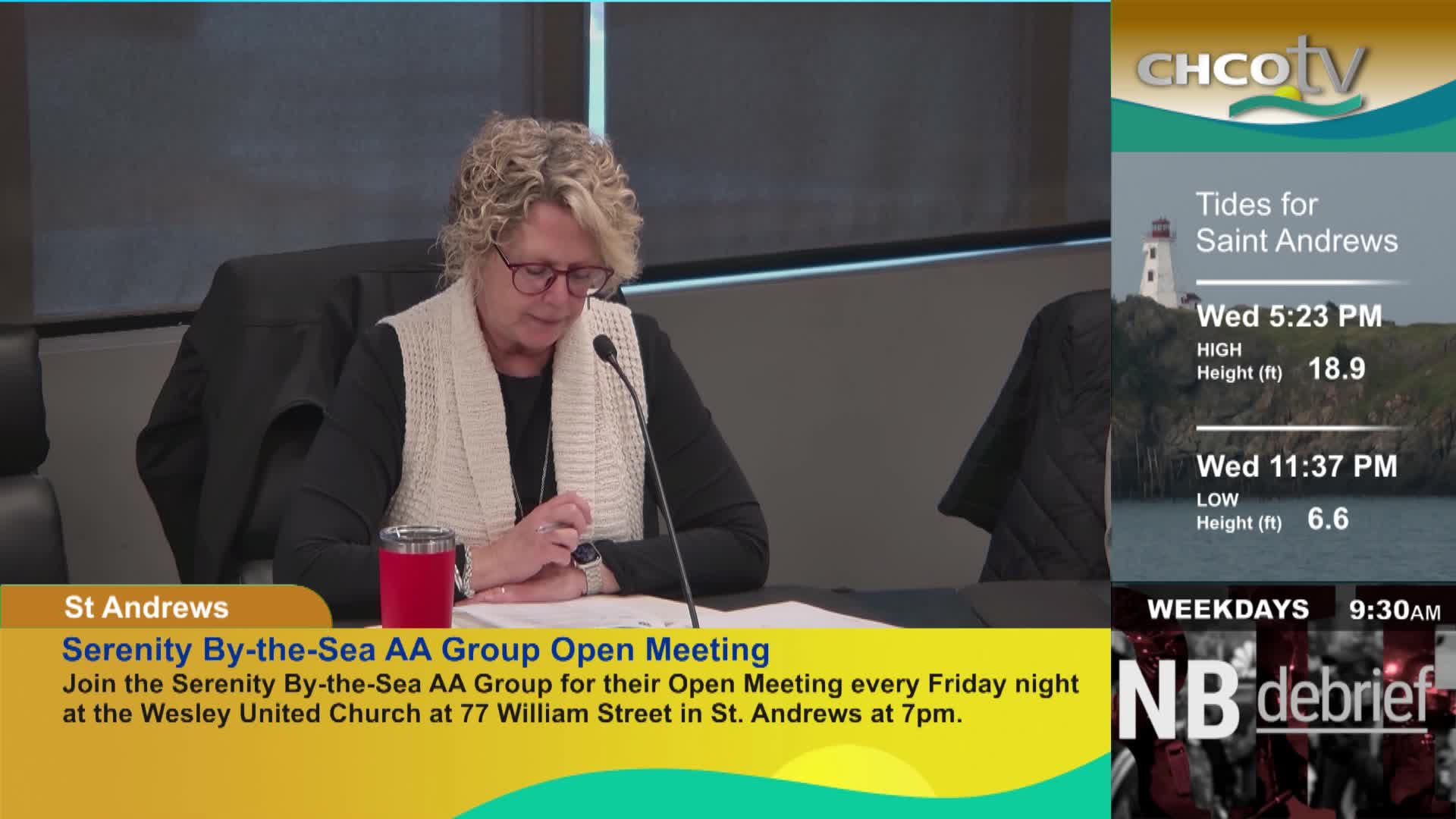 A woman with blonde, curly hair is reading from papers in front of her, a red insulated mug beside her. On the right side of the screen, CHCO-TV displays tide information for Saint Andrews.