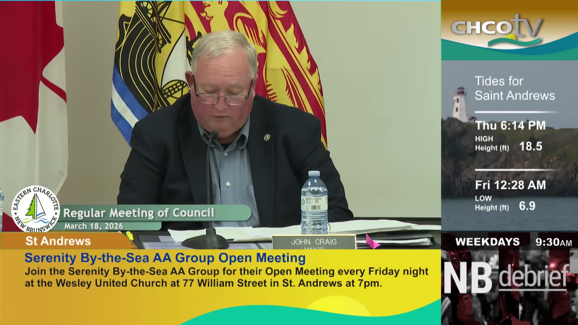 A man in a suit looks down at papers on a table. A bottle of water sits to his right.