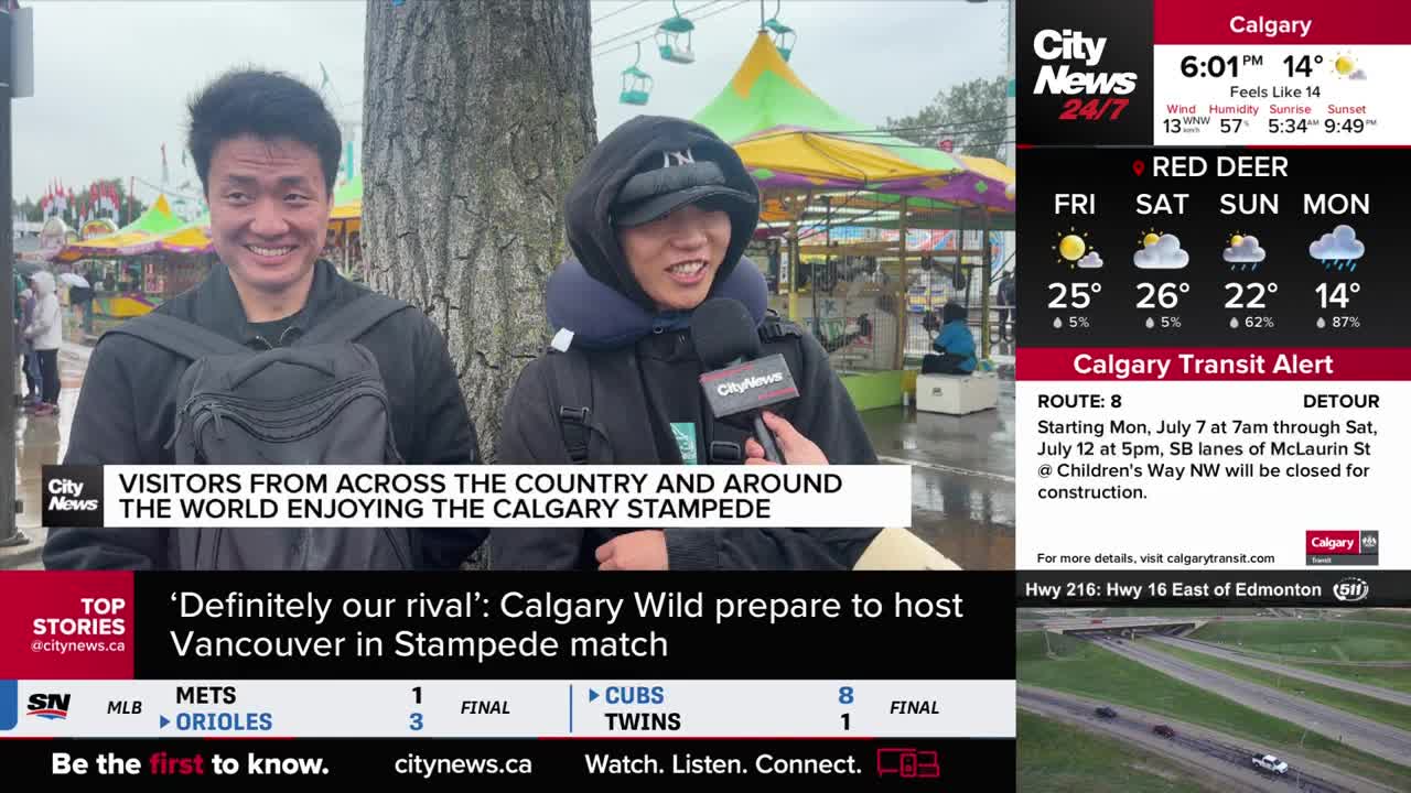 A CityNews Calgary reporter holds a microphone toward a man wearing a neck pillow and a CityNews jacket. Behind them, a fairground is visible, with colourful rides and tents in the background.
