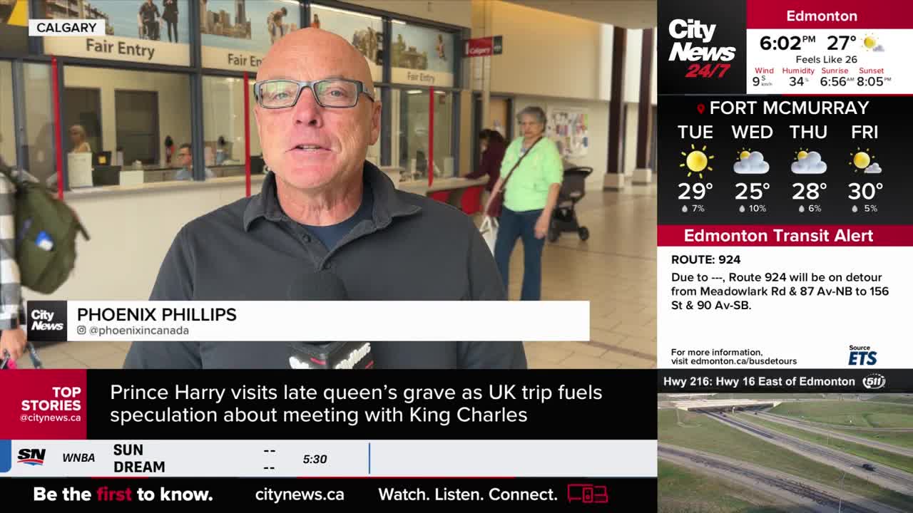 Phoenix Phillips, from CityNews Calgary, is speaking directly to the camera, a microphone in his hand. Behind him, a woman in a green shirt walks through a brightly lit hallway.
