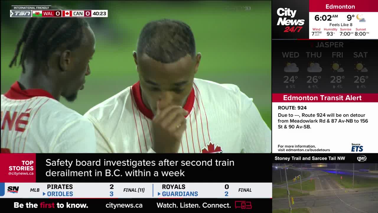Two soccer players, wearing white jerseys with the Canadian flag, are standing close together. One is touching his face, while the other looks on, the score 0-0 on the screen.
