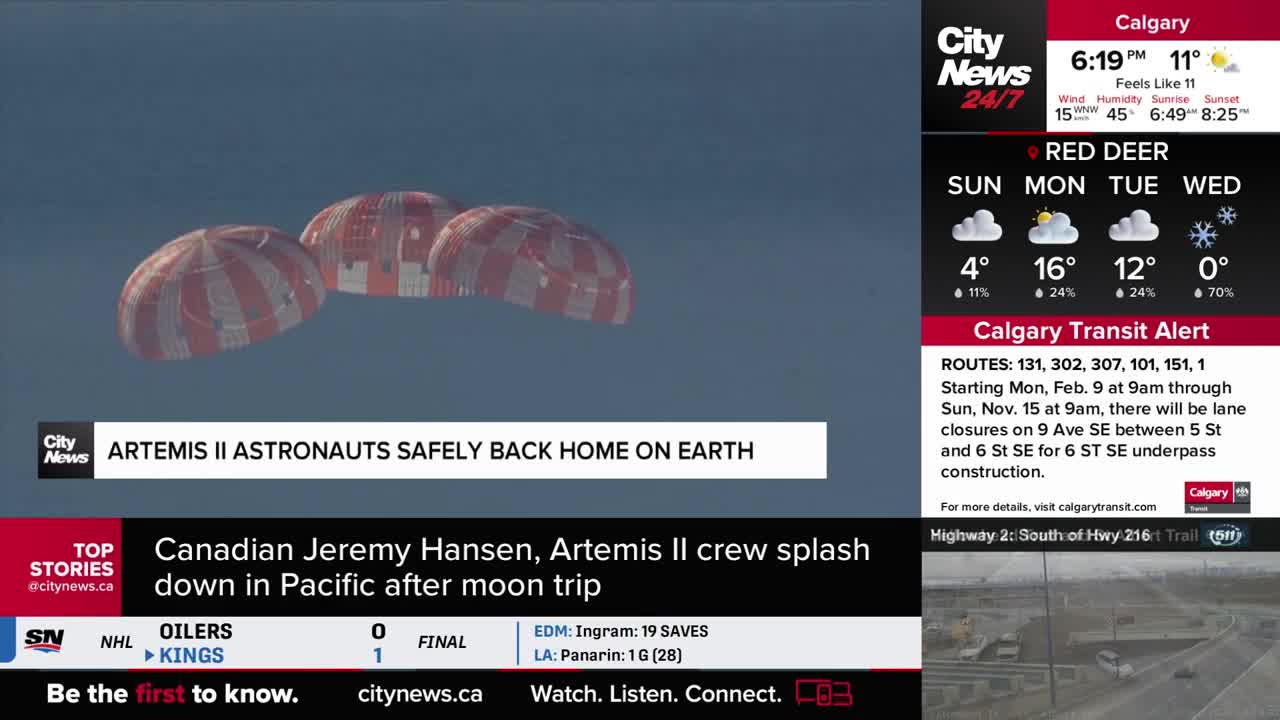 Three large parachutes, striped red and white, are deployed against a pale blue sky. This is the Artemis II crew, including Canadian Jeremy Hansen, splashing down in the Pacific after their moon trip.