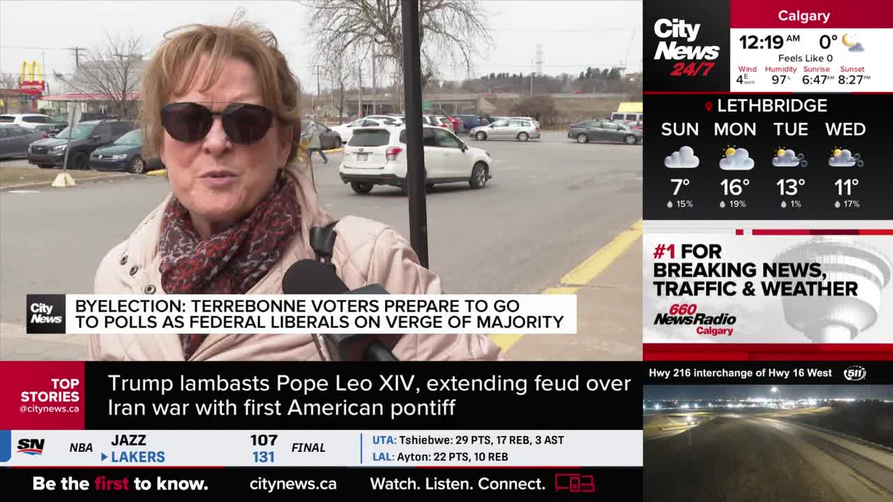A woman speaks into a microphone, her words about the Terrebonne byelection and federal Liberals likely heading for a majority. Behind her, cars move slowly through a parking lot under a grey sky.