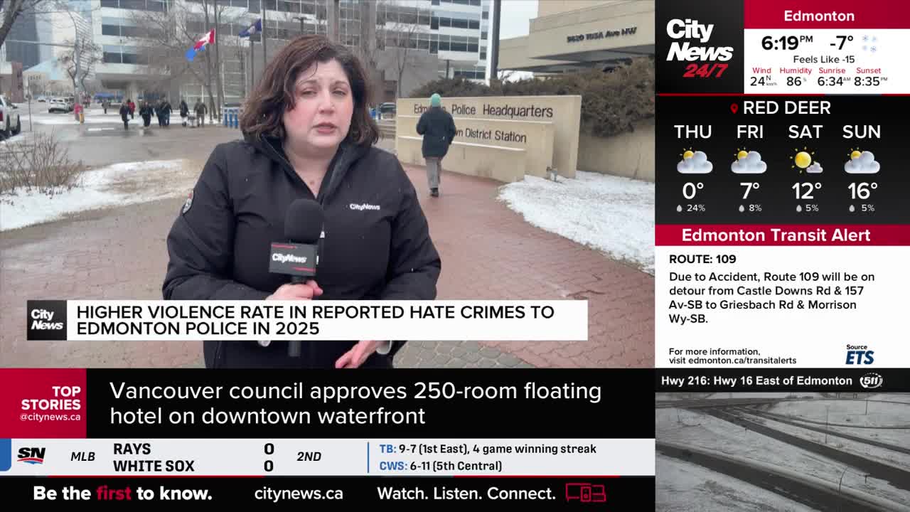 A reporter stands outside Edmonton Police Headquarters, microphone in hand. Behind her, a person in a green hat walks away from the building.