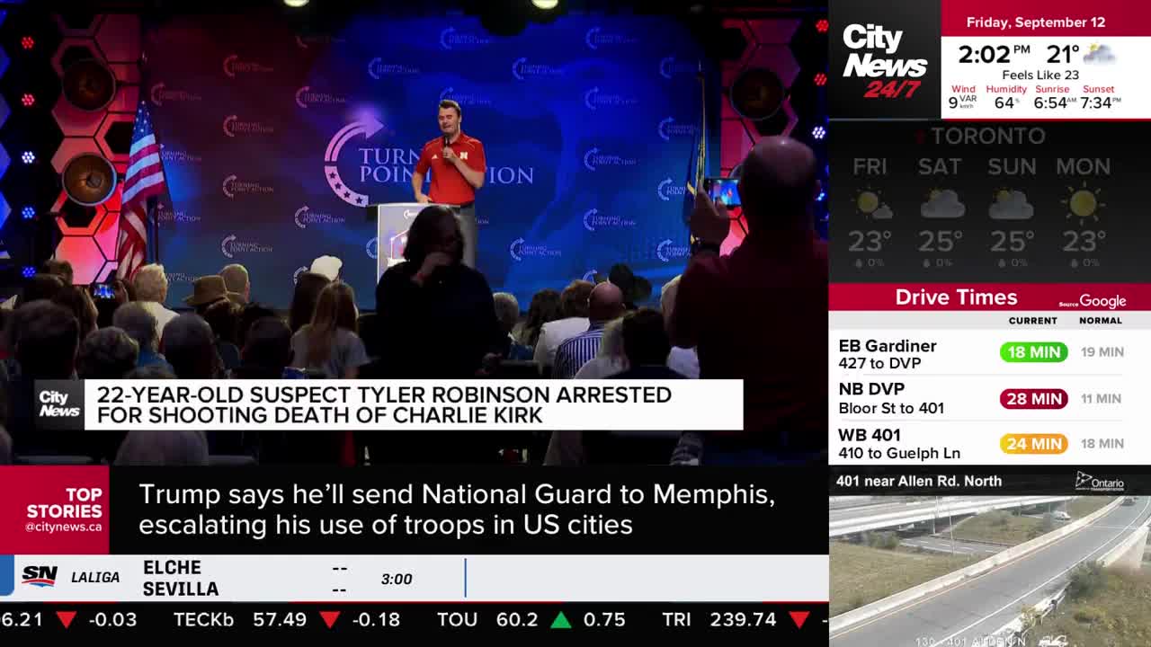 A man in a red shirt speaks at a podium, addressing a seated audience. Someone in the crowd is holding up a phone, likely recording the speaker.
