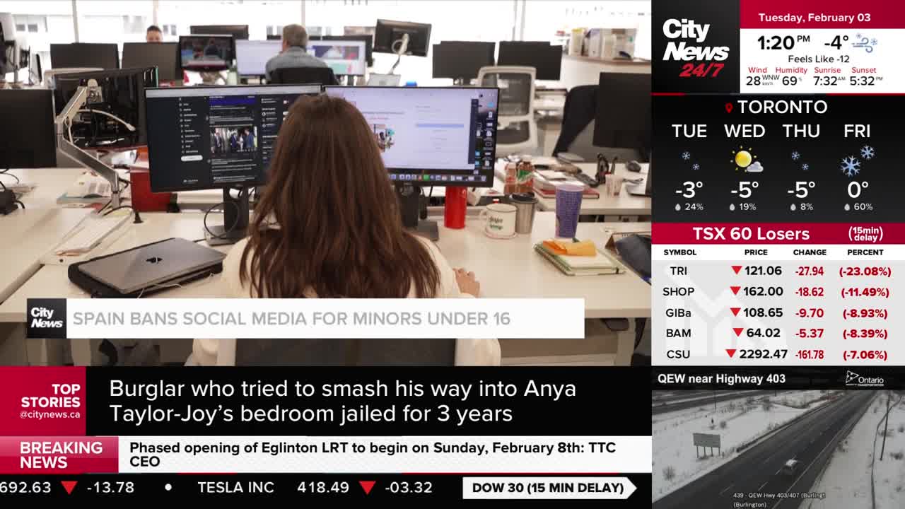 A woman with long brown hair is typing at her desk, facing away from the camera. On the screen in front of her, a CityNews headline reads "SPAIN BANS SOCIAL MEDIA FOR MINORS UNDER 16."