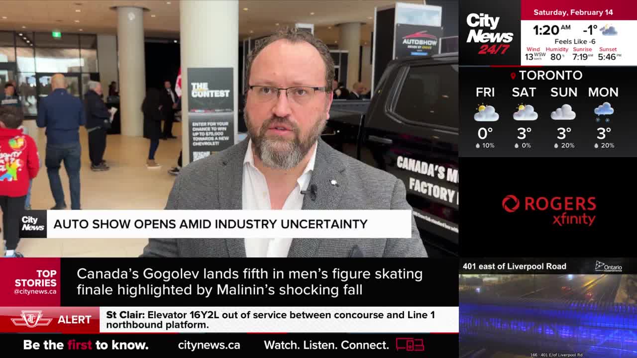 A man in a grey suit speaks directly to the camera, with a black pickup truck and an "Autoshow" sign behind him. People mill about in the background of the convention centre.