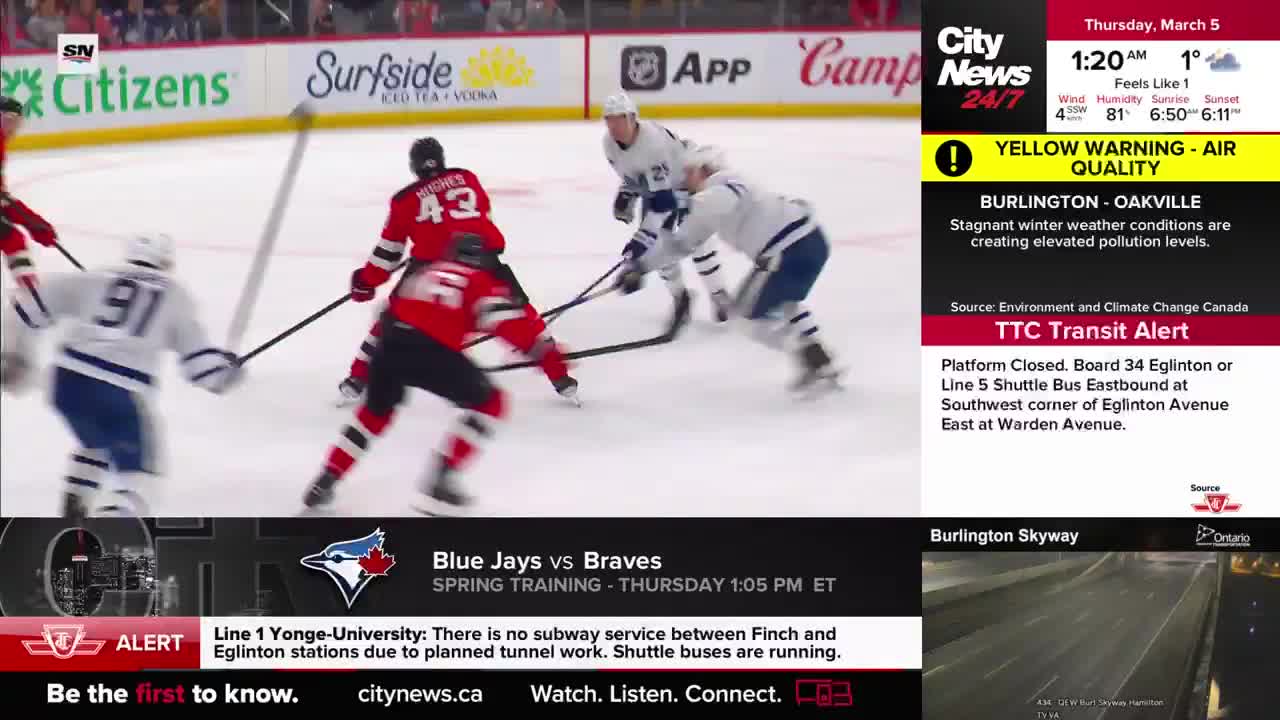Two hockey players in red jerseys battle for the puck near the boards while a player in a white and blue jersey skates towards them. A CityNews Toronto graphic displays a yellow air quality warning for Burlington and Oakville.