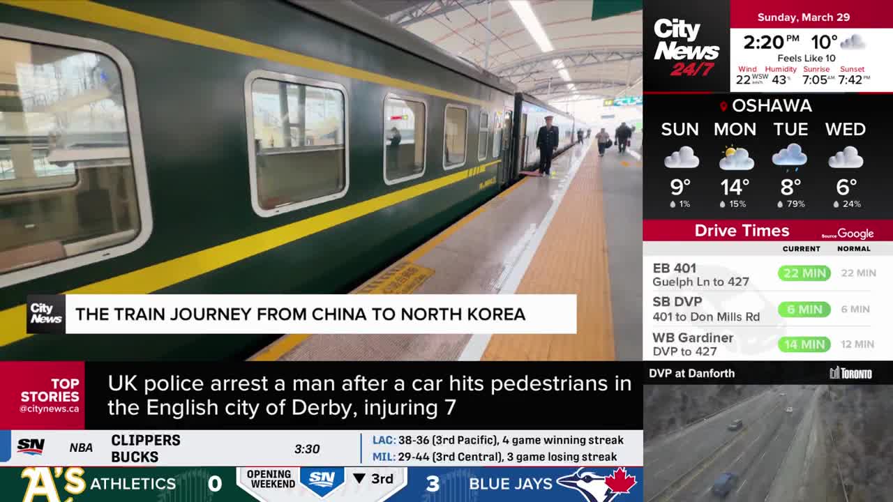 A train, painted dark green with a yellow stripe, sits at the platform. A uniformed attendant stands by the open door, while a few passengers walk away down the track.