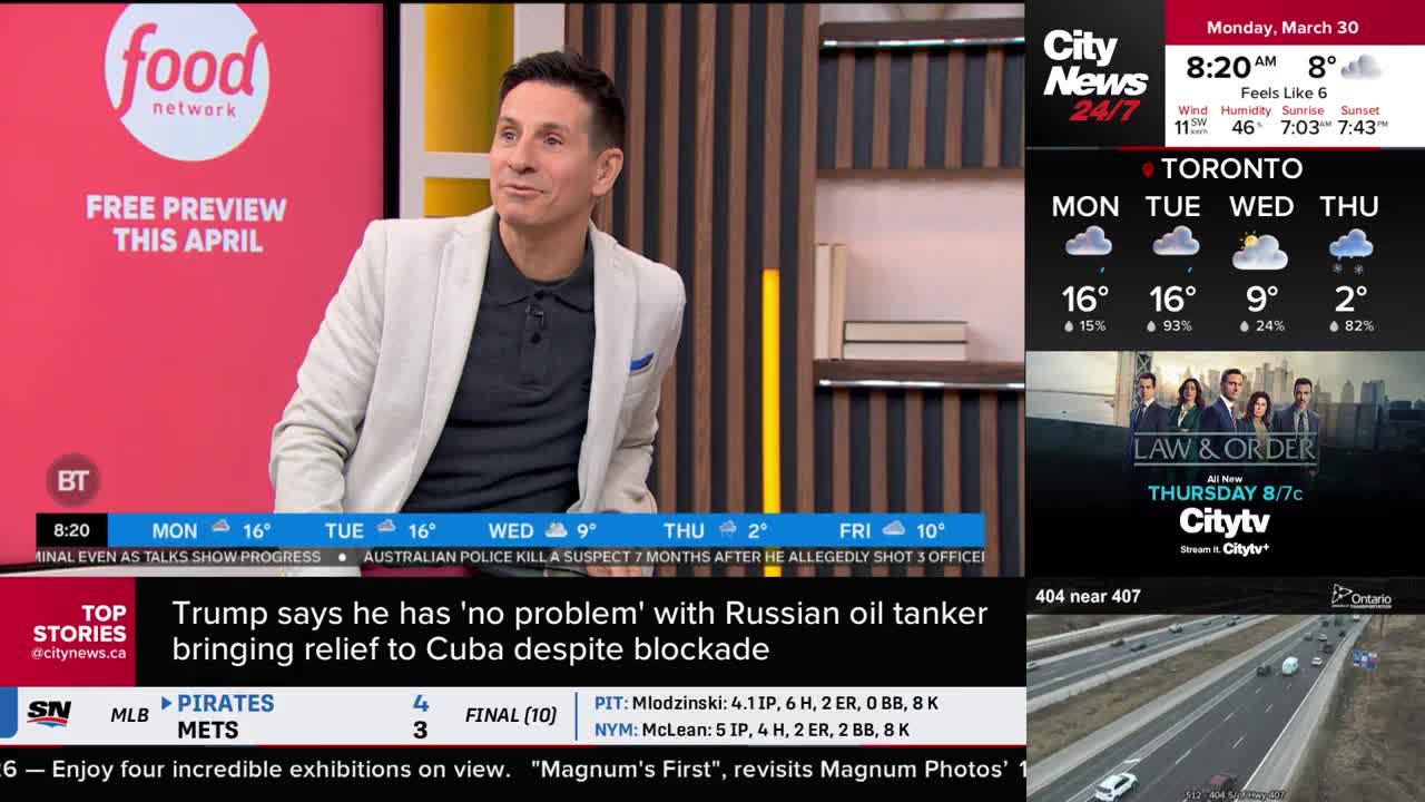 A man in a light jacket sits on a set, smiling. On the screen behind him, CityNews Toronto displays the day's weather and a preview for "Law & Order."