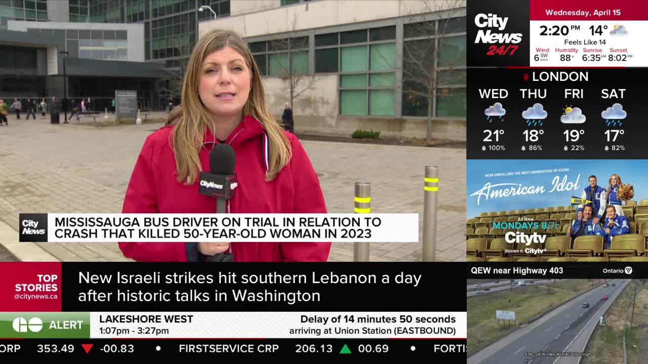 A reporter stands outside a modern building, speaking into a microphone. In the background, a few people walk by, and a highway stretches into the distance.