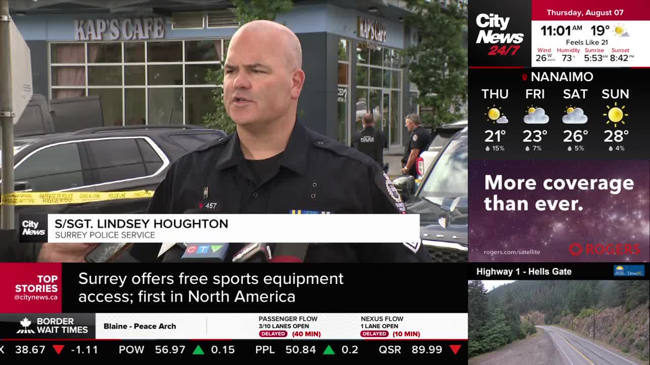 A Surrey Police officer is speaking into microphones, likely for CityNews Vancouver, with yellow tape and a black SUV in the background. Behind him, two other officers stand near a cafe entrance.
