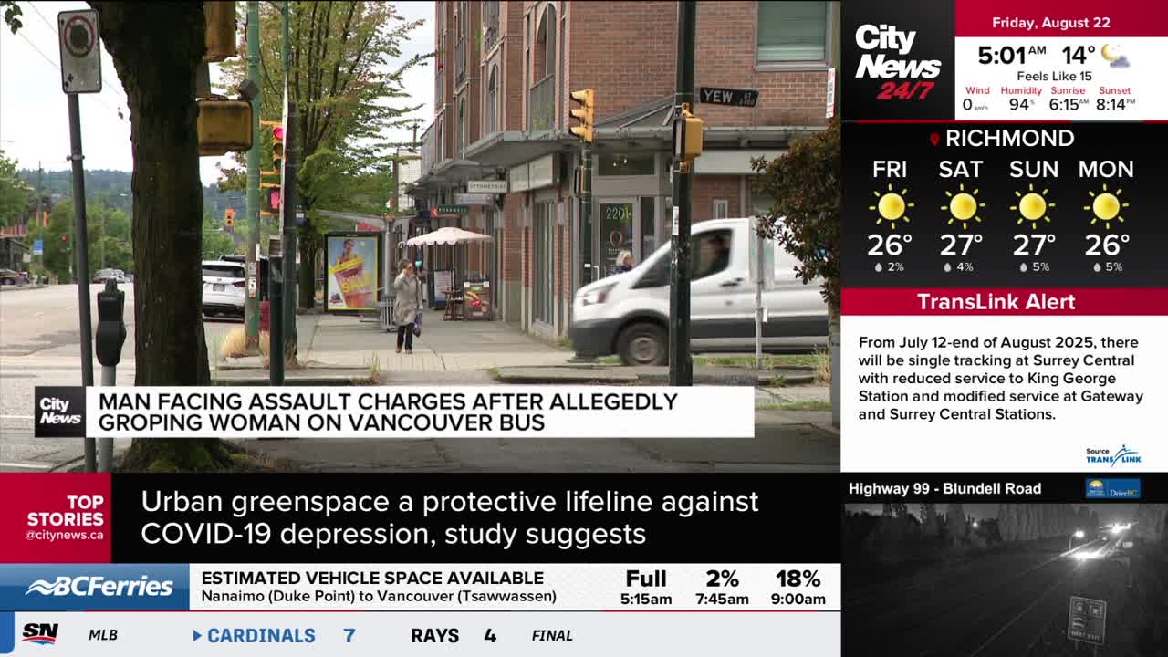A white van is turning onto a street corner in Vancouver, near a building with shops. A woman walks along the sidewalk, past a bus stop with a poster.
