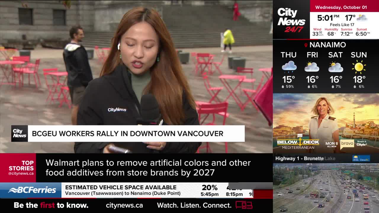 A CityNews Vancouver reporter speaks into a microphone, reporting on a BCGEU workers' rally.  Behind her, people mill about in the plaza, and a few bright pink chairs and tables sit empty.
