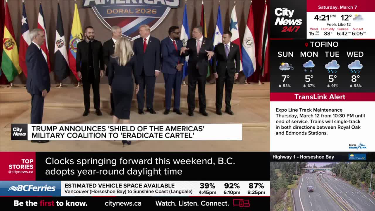 A group of men in suits stand behind a podium, some with their hands clasped. Flags of various nations are displayed behind them.
