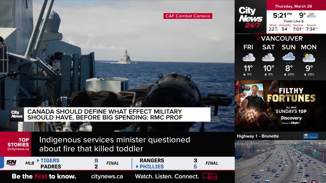 A Canadian naval vessel cuts through the blue ocean, its silhouette sharp against the horizon. In the foreground, a different ship's deck is visible, cluttered with equipment and a large, dark cylindrical object.