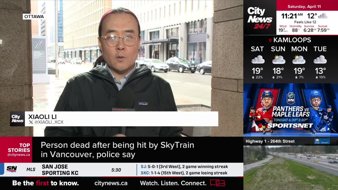 A reporter stands on a city street, speaking into a microphone. Cars move along the road behind him, and a digital display shows news headlines and weather updates.