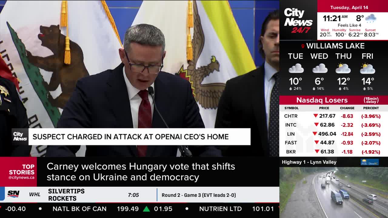 A man in a suit and glasses speaks at a podium, his gaze directed downwards. Behind him, another man in a suit stands to the side, his expression unreadable.