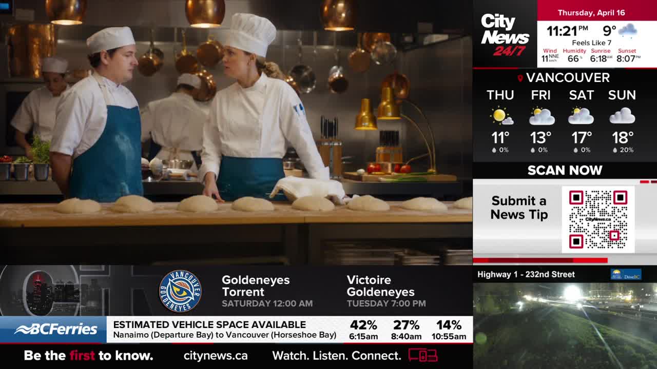 Two chefs in white uniforms and hats stand behind a counter laden with proofing dough. One chef gestures while speaking to the other, their faces illuminated by the kitchen lights.