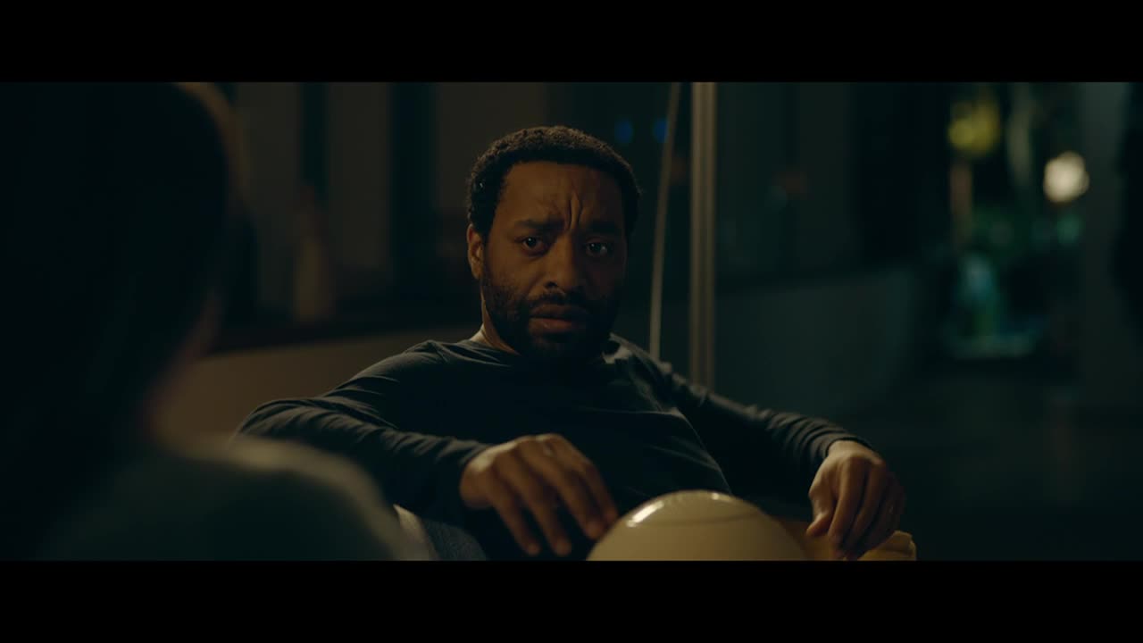 He sits, elbows resting on the chair's arms, and stares intently at someone out of frame. His expression is a mix of concern and something else, maybe weariness, as he holds a small, white object.

