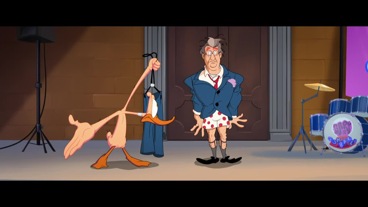 A lanky duck holds up a pair of pants while a man in a suit jacket and polka-dot underwear stands awkwardly nearby. A drum kit and a speaker stand are set up in the background.

