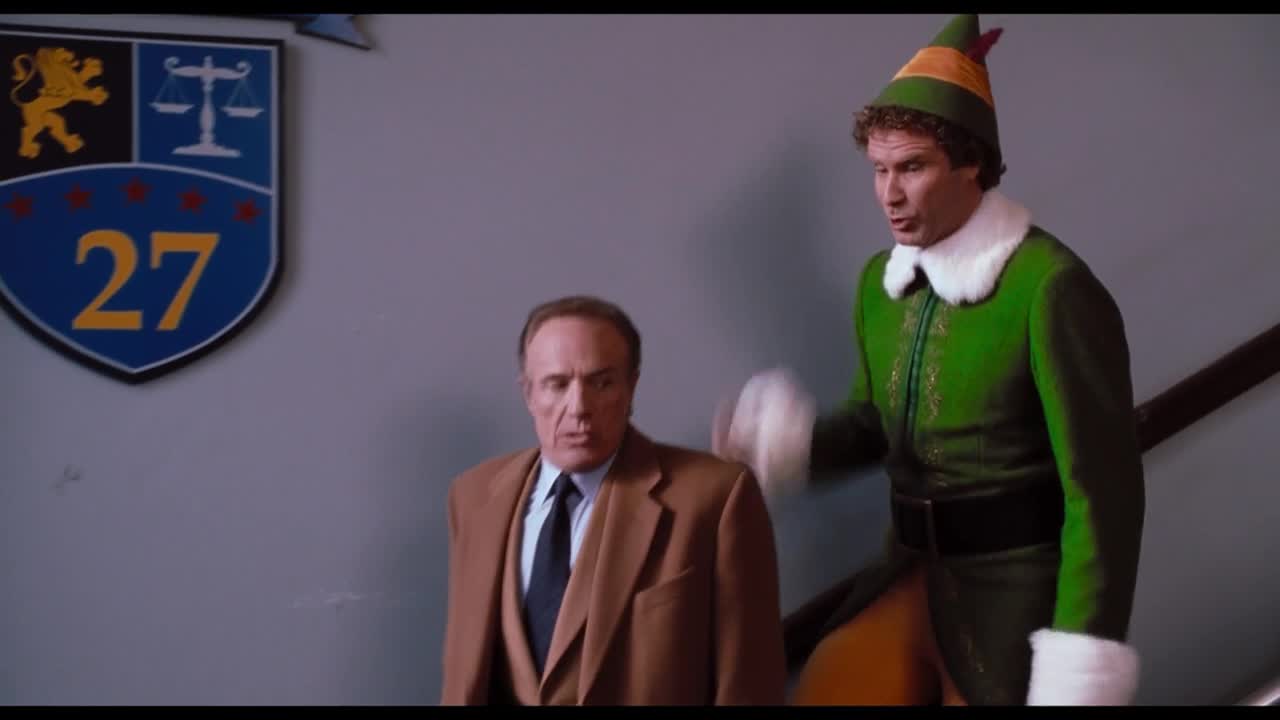 A man in a brown suit looks startled as a person in a green elf costume gestures toward him. They're descending a staircase, with a crest on the wall beside them.
