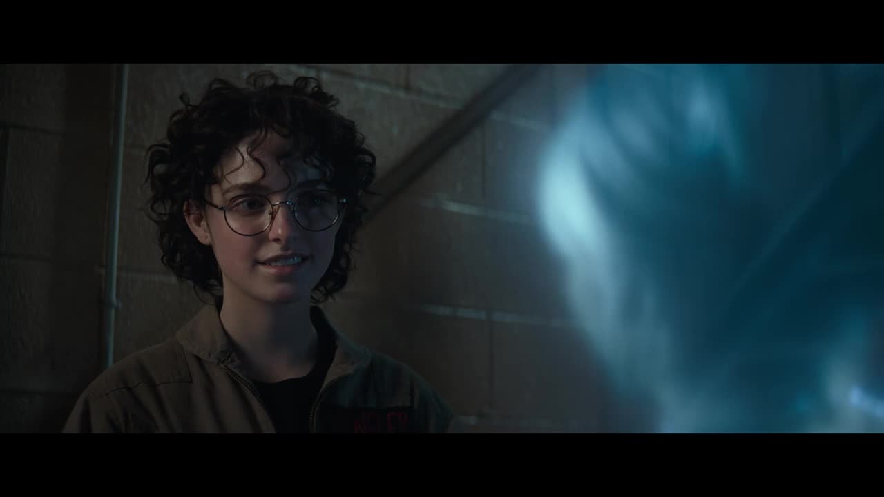 A young woman in glasses smiles, looking at something just off-screen. A translucent blue form, perhaps a ghost, hovers nearby.
