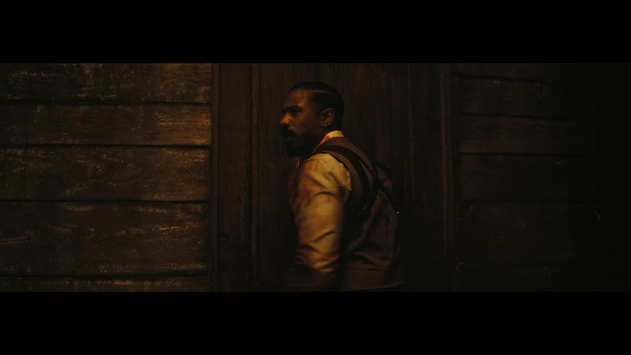A man in a vest and a backpack stands in a dimly lit space, his gaze fixed to the side. The wooden door behind him is a dark, textured backdrop.
