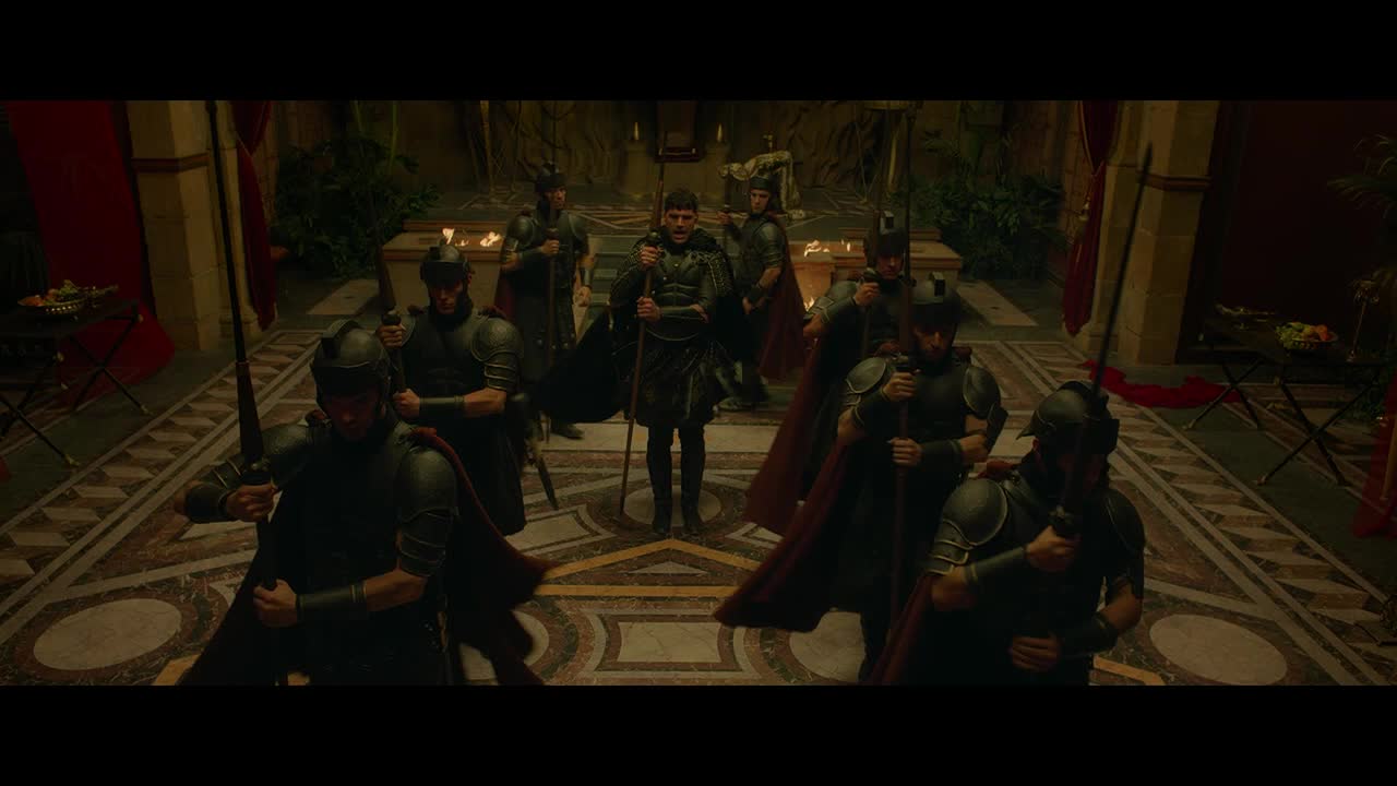 A group of armored figures, cloaked in dark red, stand at attention, holding swords. In the center, a man in similar attire, but without armor, stands with a staff.
