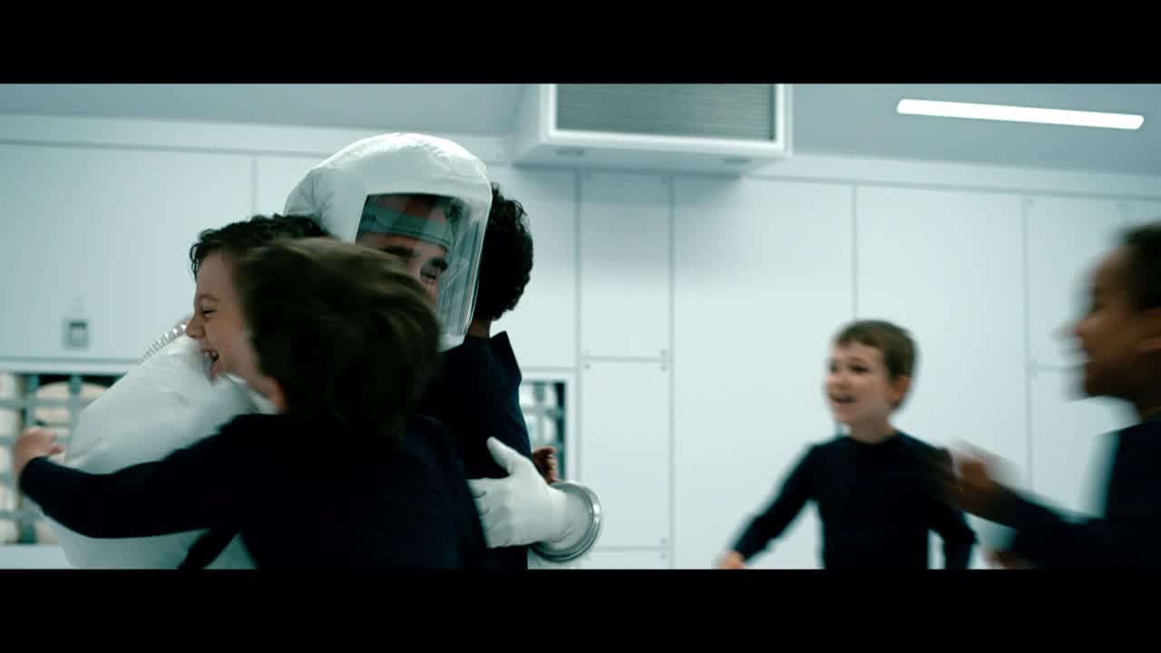A person in a white hazmat suit is embraced by two children, their faces lit up with joy. Other kids run toward them, eager to join the hug.
