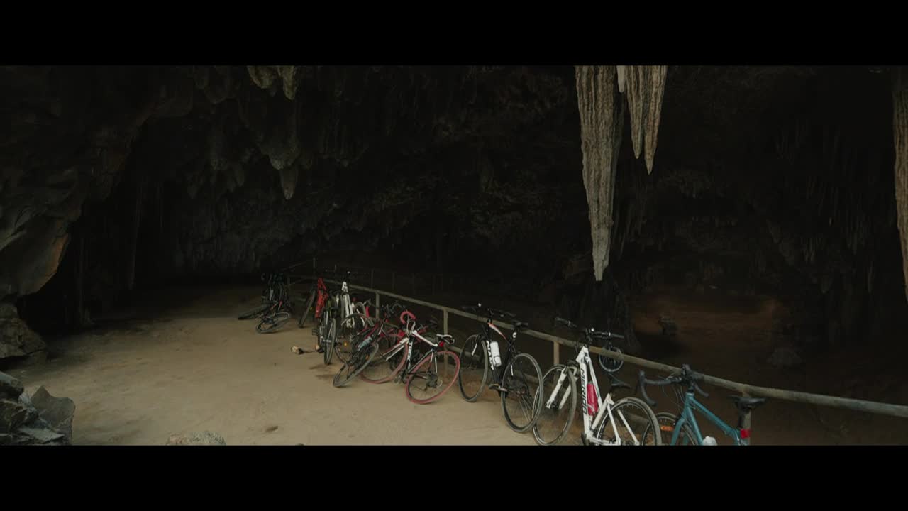 Bikes are lined up along a wooden railing inside a dark cave, their wheels still. Long, pale stalactites hang from the ceiling, illuminated by a dim light.
