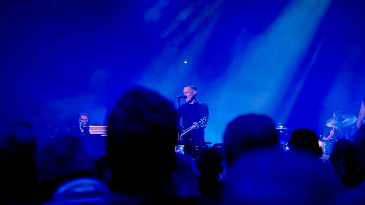 The singer, with a guitar, is in the spotlight, singing into the microphone. A keyboardist and drummer are in the background, bathed in blue light.
