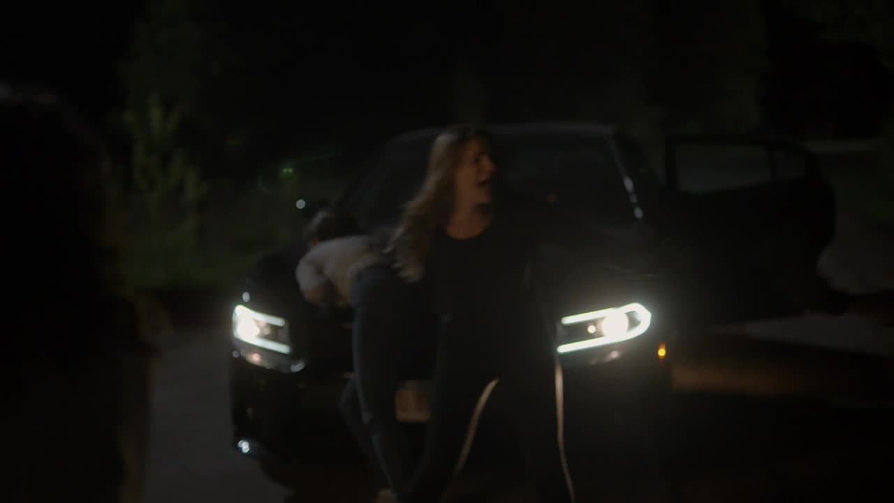 A woman in black is shouting, her face illuminated by the car's headlights. Someone is wrestling with her near the car's front.
