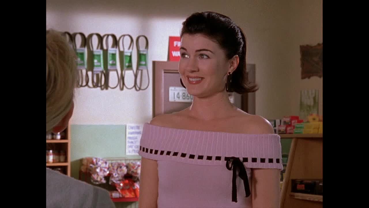 A woman in a pink off-the-shoulder top smiles, looking at someone off-screen. Behind her, a shelf displays car parts and a sign that reads "FREE WATER."
