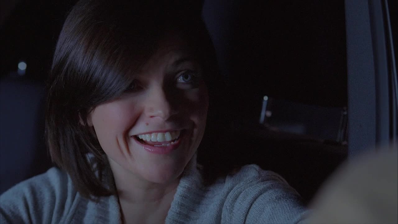 A woman in a car smiles, her eyes crinkling at the corners. The interior is dark, but her face is illuminated by an unseen light source.
