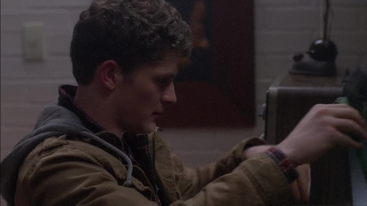 A young man in a hooded jacket is focused on something out of frame, his hands gently moving. The dim lighting and the vintage radio suggest a scene from a Canadian drama on Crave 2.
