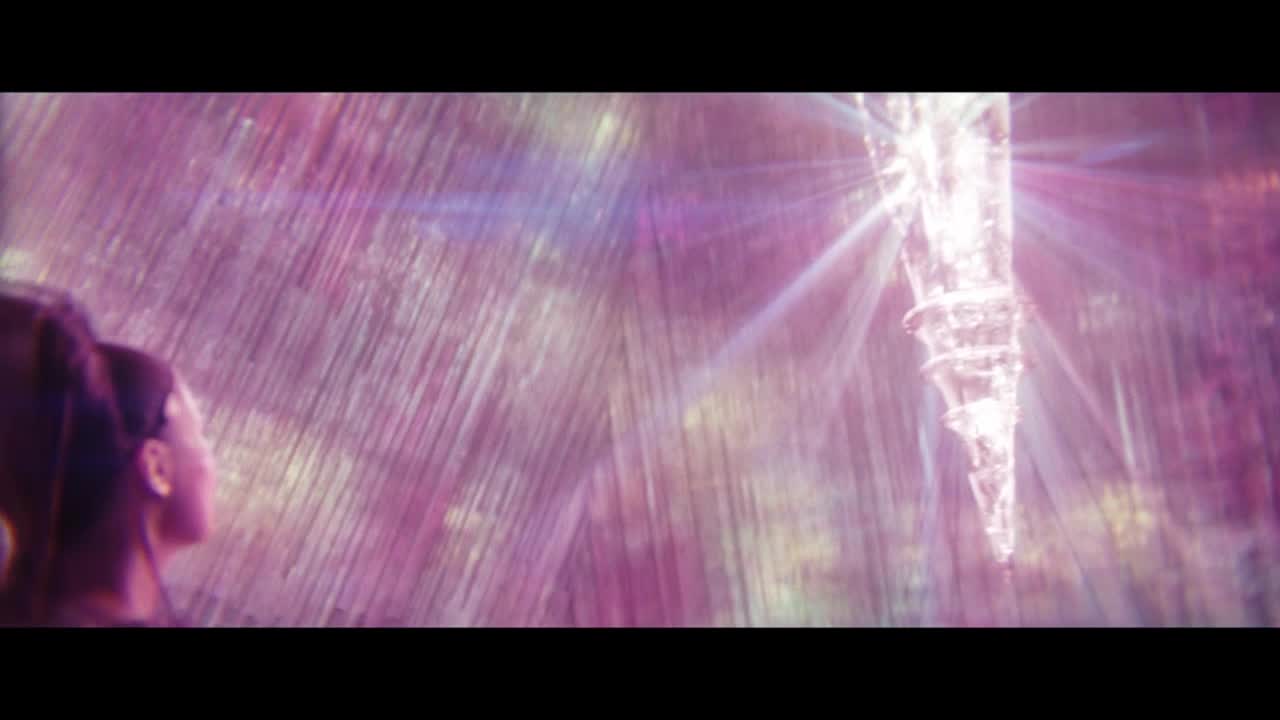 A woman gazes upward as a glowing, crystalline structure descends. Light streaks across the frame, creating a shimmering, ethereal effect.
