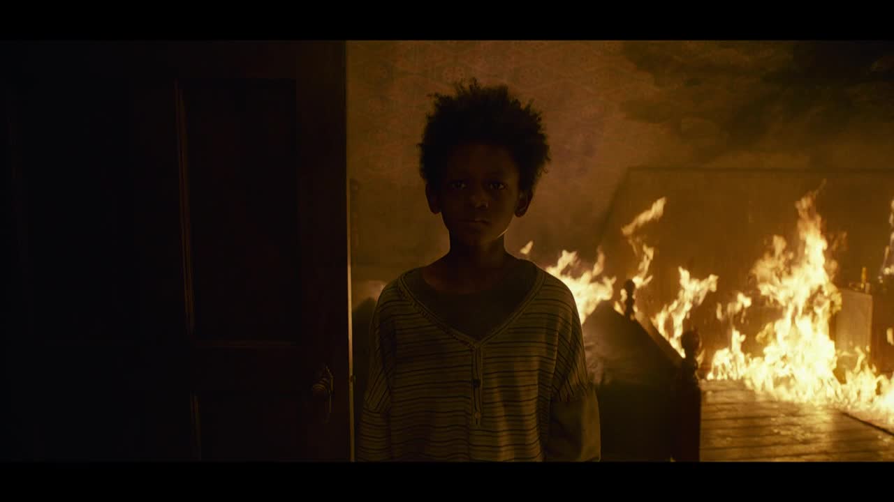 A young person stands in a doorway, their gaze steady as flames consume the room behind them. The fire licks at the furniture, casting a warm glow on the child's face.
