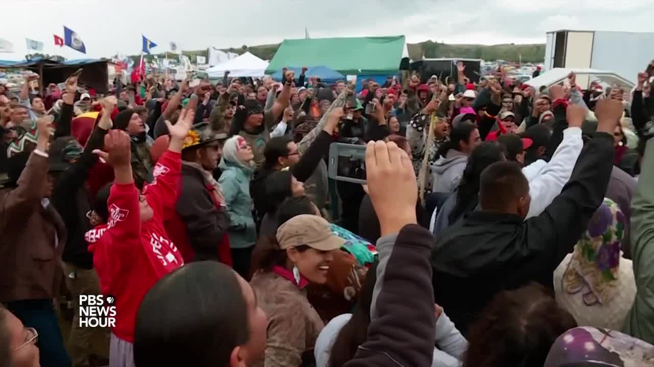 A large crowd of people is gathered, many with their arms raised in the air. Flags and tents are visible in the background, as people cheer and record the scene on their phones.
