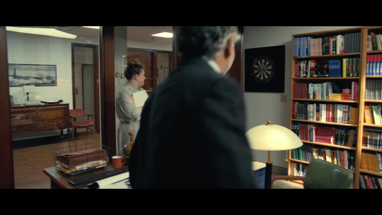 A man in a dark suit walks toward the camera, his back to us. In the background, a woman stands in an office doorway, holding a stack of papers.
