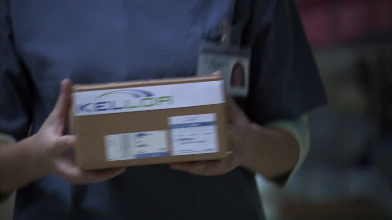 A person in scrubs holds a cardboard box labeled "KELLOR" in front of them. The box is being carried, likely in a hospital setting, as seen in a Canadian Crave 2 production.
