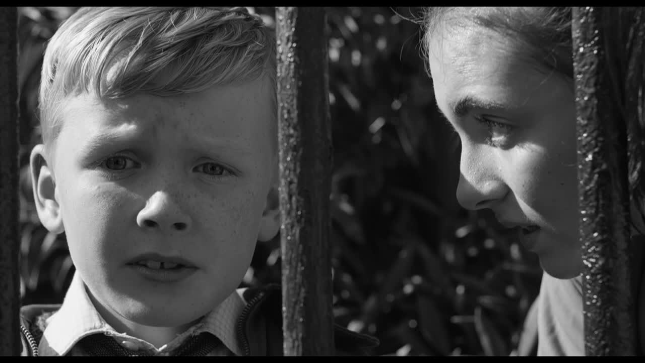 A young boy stares directly ahead, his face framed by the bars of a gate. Beside him, a girl leans in, her gaze directed toward him.

