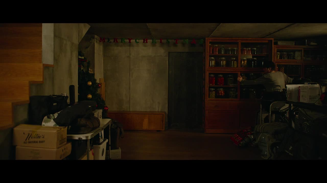 A man is rummaging through a cabinet filled with jars, his back to me. The basement is cluttered, with a Christmas tree and boxes stacked near the stairs.

