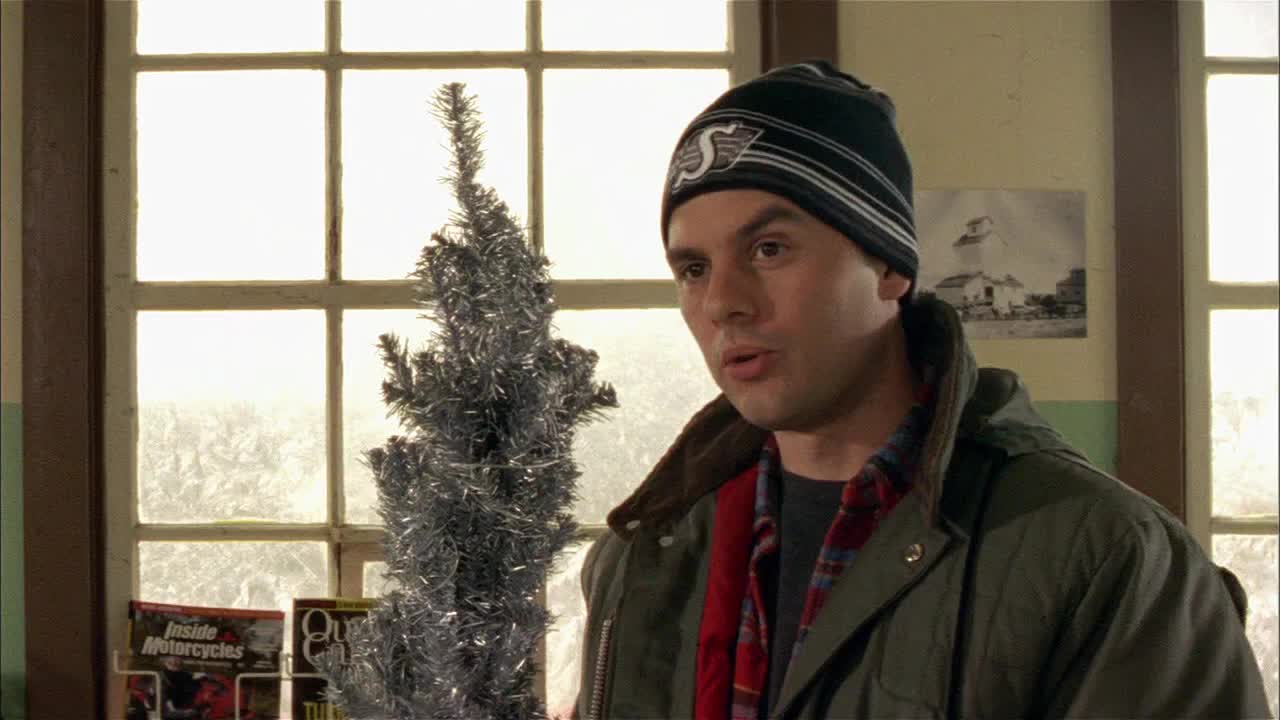 A man in a toque and jacket gestures with his hands, speaking near a small, silver Christmas tree. Behind him, a faded black and white photo hangs on the wall, and a window lets in the daylight.
