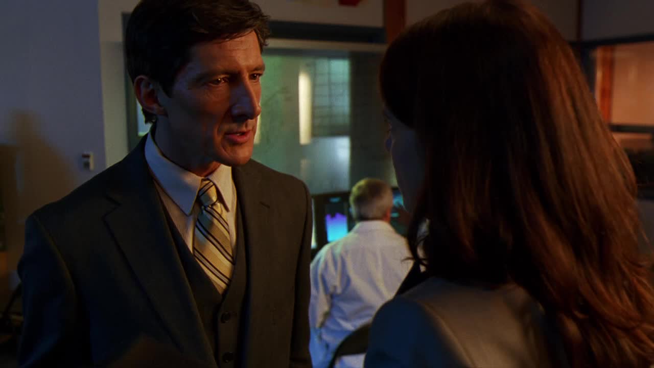 The man in the suit is speaking intently, his gaze fixed on the woman in front of him. Behind them, a man in a white shirt works at a computer in a brightly lit room.
