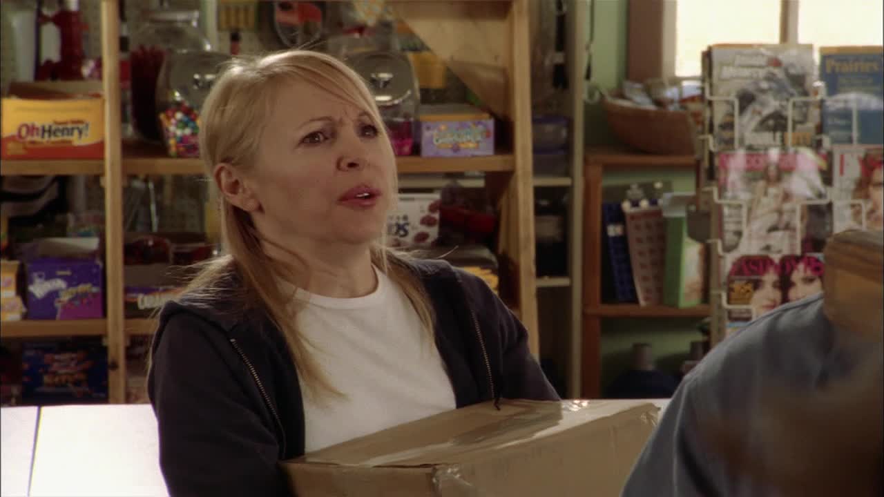 A woman in a white t-shirt and a dark jacket holds a cardboard box, her expression a mix of concern and surprise. Behind her, shelves are stocked with candy and magazines, a classic Canadian convenience store scene.
