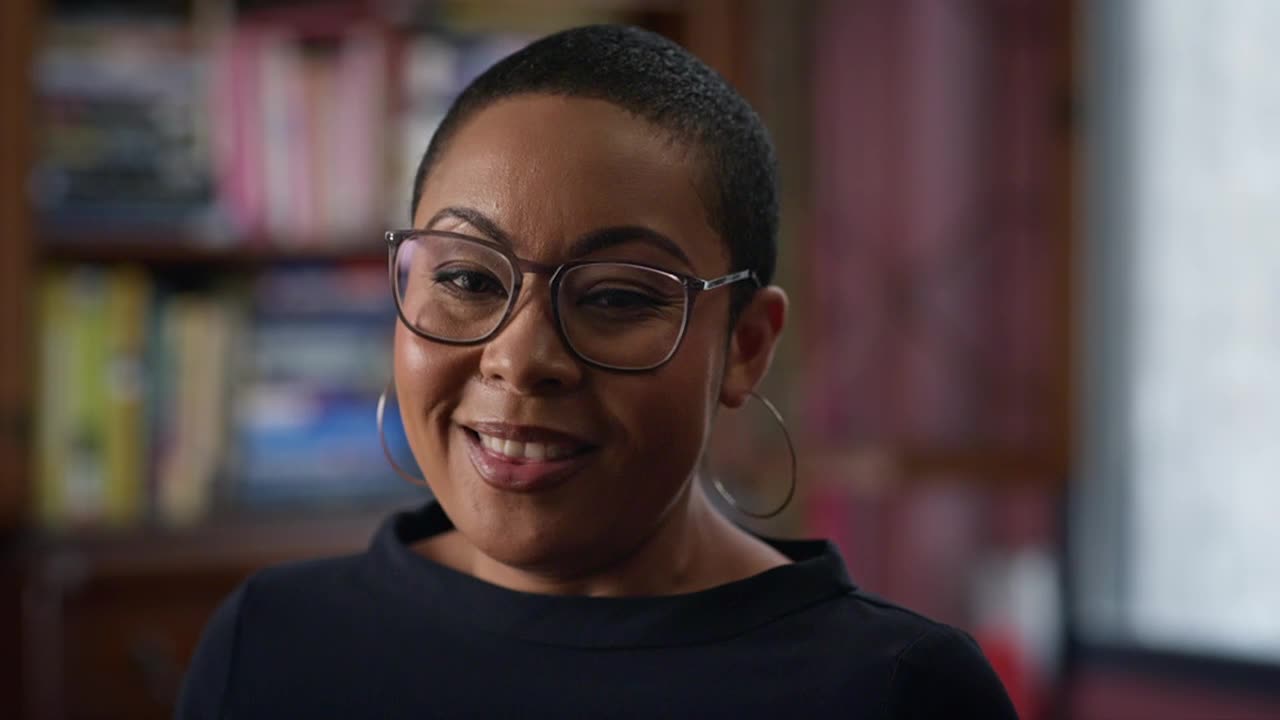 A woman wearing glasses and hoop earrings smiles directly at the camera. Behind her, a bookshelf filled with books is slightly out of focus.
